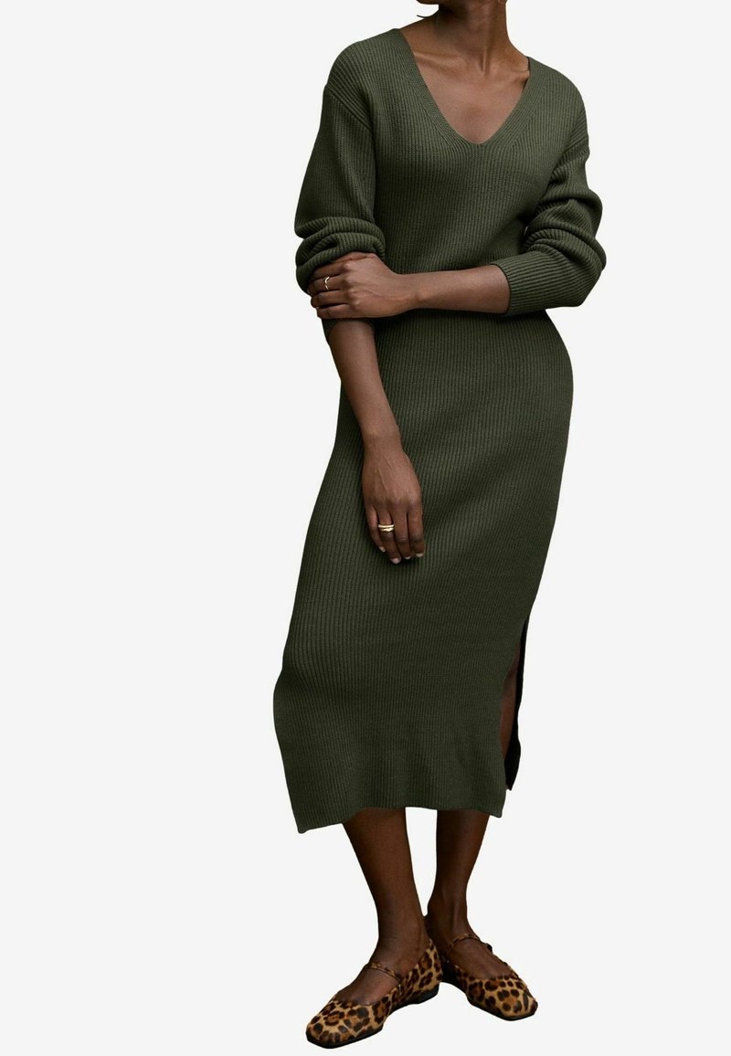 Femme portant une longue robe en maille côtelée vert olive avec un col en V et des ballerines à imprimé léopard, debout les bras croisés.