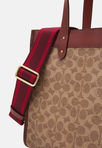 Tan canvas tote bag med sammenflettet mønster; brune læderdetaljer; justerbar marsala-farvet rem med røde striber og guld spænde.