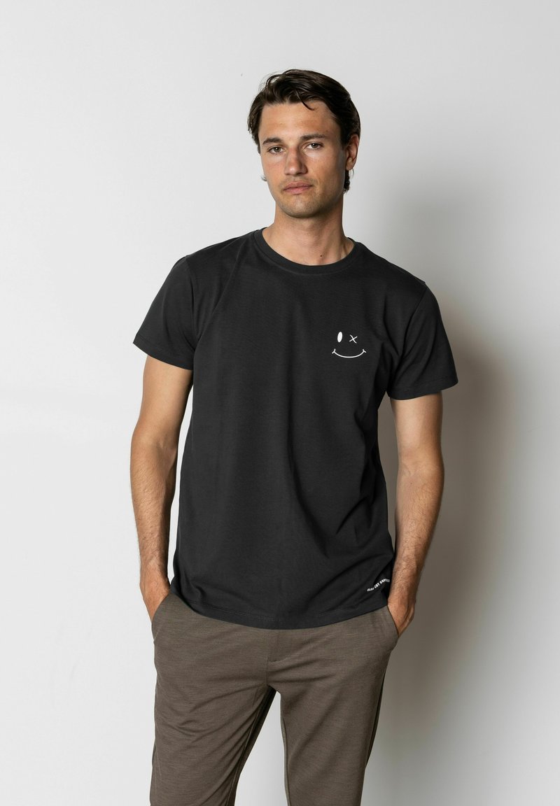 T-shirt en coton noir à manches courtes et col rond. Présente un graphique blanc d'un visage souriant sur le côté gauche de la poitrine.