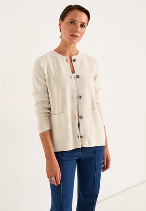 Cremefarbener Strickcardigan mit Rundhalsausschnitt, Knopfverschluss vorne, zwei Vorder-taschen und gerippten Bündchen. Getragen über einem weißen Oberteil und blauen Hosen.