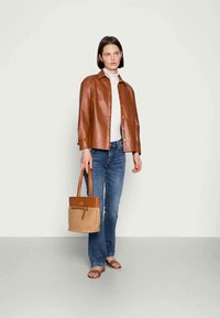 Brown leather shirt jacket, white turtleneck, blue jeans, and a tan woven handbag. Worn with flat sandals, standing against a neutral backdrop.
