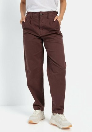 Personne portant un pantalon marron à jambes droites avec des ourlets élastiques, les mains dans les poches, associé à des baskets blanches sur un fond uni.