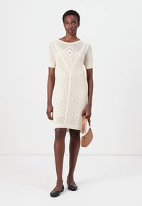 Cream-colored knitted dress with a geometric diamond pattern, short sleeves, a fitted silhouette, and ribbed hem; paired with a small tan handbag.