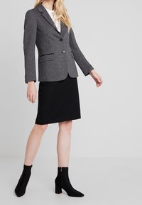 Blazer grigio a scacchi con due tasche frontali, chiusura a un bottone, abbinato a una gonna nera al ginocchio e stivali alla caviglia neri.