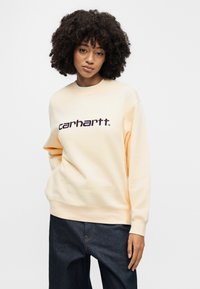 Νεαρή γυναίκα με σγουρά μαλλιά, ντυμένη με ένα κρεμ φούτερ Carhartt και σκούρο τζιν, ποζάροντας μπροστά σε απλό λευκό φόντο.