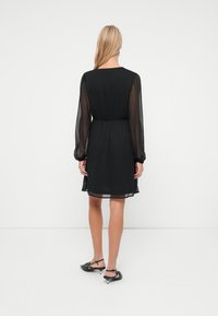 Nainen, jolla on pitkät vaaleat hiukset, pukeutuneena polvipituiseen mustaan mekkoon, jossa on läpinäkyvät pitkät hihat ja mustat kitten-heel slingback-kengät, seisoo selkä kameralle.