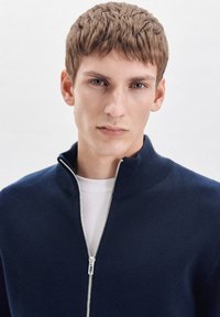 Marnet zip-up trui met hoge kraag, gebreide textuur en zilveren ritssluiting, gedragen over een wit T-shirt. Heeft een getailleerd ontwerp.
