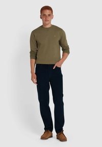 Olijfgroene sweatshirt met lange mouwen, ronde hals en een klein logo. Gecombineerd met donkerblauwe broek en bruine schoenen, in een staande pose.