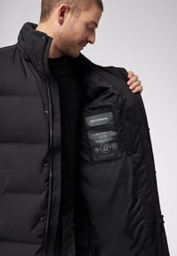 Giacca a vento nera con alto colletto, chiusura zip e tasche laterali doppie. L'interno presenta un'etichetta di cura e simboli di cura su un tessuto opaco.