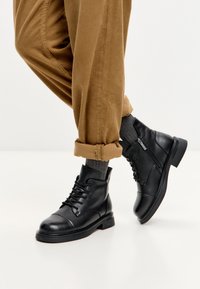 Persona con pantalones de pana marrón arremangados, calcetines grises y botas de cuero negras con cordones y cremallera lateral.