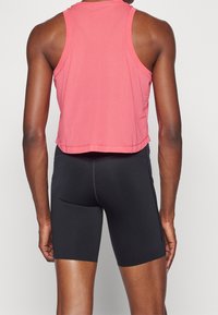 Personne portant un haut de sport rose sans manches et un cuissard de cyclisme noir ajusté, vue de dos sur un fond uni.