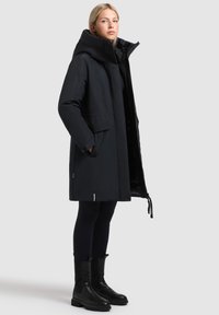 Palton parka negru cu guler înalt, siluetă largă și închidere cu fermoar. Caracteristici include buzunare laterale mari și șnururi la tiv.