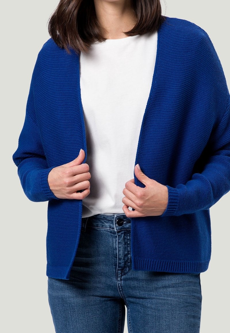 Femme portant un cardigan bleu tricoté par-dessus une chemise blanche et un jean bleu, tenant les bords du cardigan avec les deux mains.