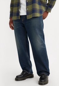 Lediga, mörkblå denimjeans med urblekta detaljer och raka ben. Kombinerade med svarta läderskor och en olivgrön flanellskjorta.