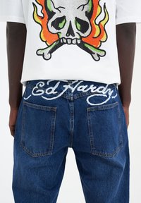 Camiseta blanca con un diseño de calavera y llamas; vaqueros azules con "Ed Hardy" bordado en blanco en la cintura, con bolsillos tradicionales.