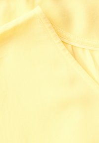 Žlutá tkanina s hladkým povrchem a výstřihem ve tvaru V. Švy jsou viditelné podél okrajů, což zdůrazňuje jednoduchost designu.