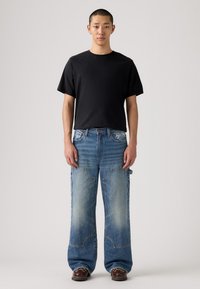 Camiseta de algodón negra combinada con unos pantalones vaqueros de denim azul anchos y desgastados que presentan grandes bolsillos, decoloración y un corte único. Zapatos de cuero marrón.