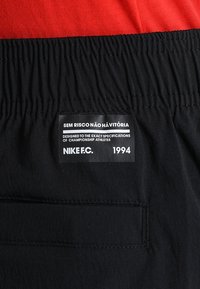 Sort taljebånd med et broderet mærke, der står "Sem Risco Não Há Vitória," "Designet til mesterskabsatleter," og "NIKE F.C. 1994" under et rødt stof.