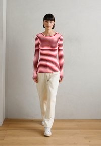 Red and white striped long-sleeve shirt paired with cream-colored pants, worn with white sneakers, on a wooden floor with a plain background.