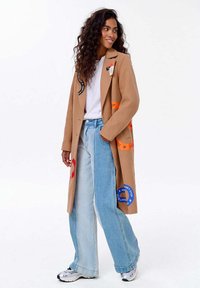 Žena v béžovém kabátu s barevnými nášivkami, bílé tričko, dvoubarevné široké džíny a bílé tenisky, stojící s jednou rukou v kapse.