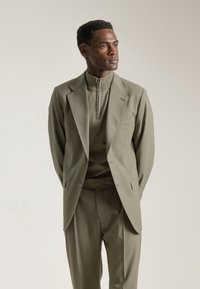 Traje verde oliva con chaqueta estructurada y pantalones a juego, que presenta una blusa de punto de alto cuello. Textura suave y ajuste a medida.