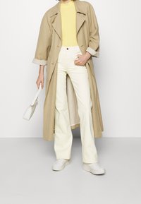 Beige trenchcoat, gul skjorte, hvide højtskårne bukser, hvide sneakers og en lille hvid håndtaske. Enkel design, bløde teksturer.