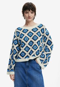 Pull en crochet avec des motifs en diamants bleu et noir, base blanche, manches larges et coupe décontractée, accompagné d'un bas en denim bleu.