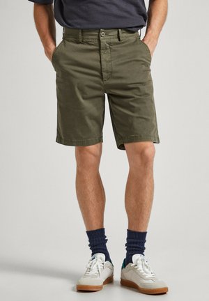 Pantalones cortos de algodón verde oliva con múltiples bolsillos y cierre de botón, con un corte recto y un ajuste relajado. Llevados con zapatillas blancas y calcetines azul marino.