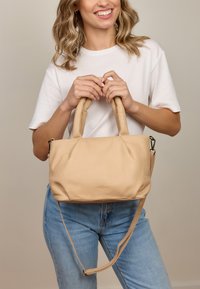 Uśmiechnięta kobieta trzyma beżową skórzaną torebkę z wyściełanymi uchwytami i odczepianym paskiem na ramię, ubrana w białą bluzkę i niebieskie dżinsy.