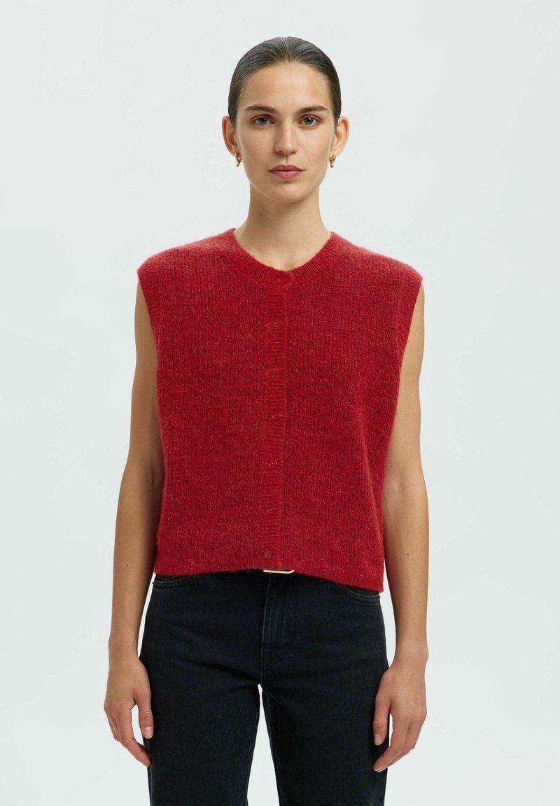 Top tricot sans manches rouge avec un encolure ronde et une fermeture à boutons sur le devant. Texture tissée et coupe décontractée, associé à un jean sombre.