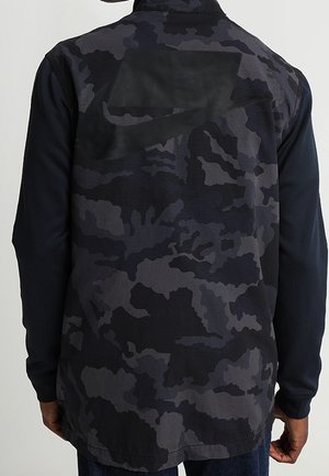 Homme portant une veste à motif camouflage noir et gris foncé vu de dos.