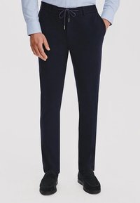 Granatowe spodnie wykonane z miękkiej tkaniny, z elastycznym paskiem w talii, dopasowanym krojem i kieszeniami po bokach. Noszone z czarnymi butami.