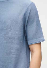 Κοντινή λήψη φωτεινού γαλάζιου υφασμάτινου κοντομάνικου πουκάμισου στον άνω βραχίονα και τον ώμο ενός ατόμου, πάνω σε απλό φόντο.