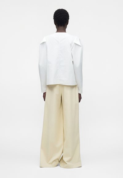 Personne aux cheveux courts et bouclés, portant un haut blanc à manches longues et un pantalon beige large, debout de dos face au spectateur sur un fond blanc.