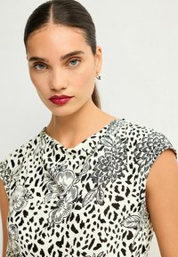 La blouse a maniche corte presenta uno sfondo crema con motivi floreali neri e stampa leopardata, scollo drappeggiato e una texture di tessuto morbida.