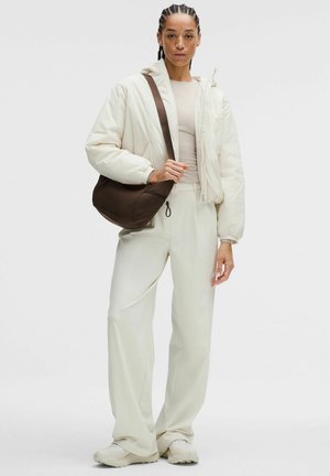 Donna con capelli intrecciati che indossa una giacca a vento bianca, pantaloni abbinati, sneaker bianche e porta una borsa a tracolla marrone scuro.