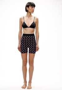 Kobieta z krótkimi ciemnymi włosami, ubrana w czarny trójkątny biustonosz i czarne szorty z wysokim stanem ozdobione różowymi wzorami półksiężyców, stojąca boso na białym tle.
