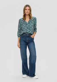 Bluse mit Blumenmuster in Blau, Grün und Weiß, kombiniert mit hochtaillierten, locker sitzenden, dunkelblauen Jeans und weißen Turnschuhen.