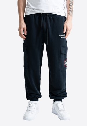 Homme portant un pantalon de survêtement cargo noir avec le logo « Ocean Pacific » et des baskets blanches, debout sur un fond blanc.