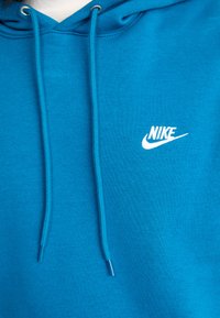 Šviesiai mėlyna Nike džemperis su balta logotipu ant krūtinės ir atitinkamomis mėlynomis raištelėmis, kabančiomis nuo gobtuvo.