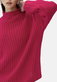 Roze gebreide trui met een hoge hals en geribbelde textuur, met verlaagde schouders en brede mouwen, die een zachte, gezellige uitstraling heeft.