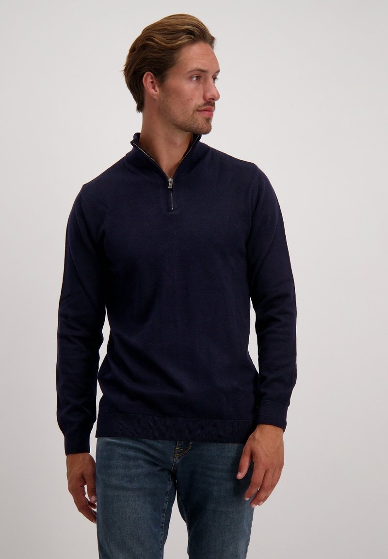 Marineblauer Pullover mit halbem Reißverschluss, gestrickter Textur und gerippten Bündchen und Saum, kombiniert mit blauen Jeans.