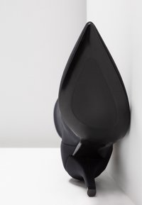 Chaussure à talon haut noire à bout pointu, positionnée avec la semelle face à l'avant sur un fond blanc.