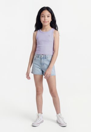 Νεαρή κοπέλα που στέκεται φορώντας αμάνικο τοπ σε ανοιχτό μοβ χρώμα, ανοιχτόχρωμα τζιν σορτς και λευκά ψηλά αθλητικά παπούτσια, με φόντο λευκό.