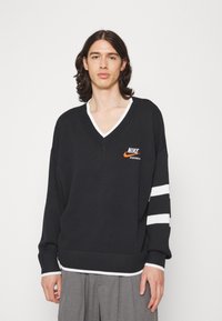 Čierny pletený sveter s výstrihom do V a bielym lemovaním, s oranžovým logom Nike a bielymi pruhovanými akcentmi na rukávoch. Voľný strih.