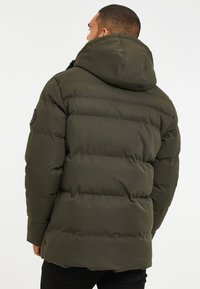 Grønne puffer-jakke med hætte, der har en quiltet design og synlige sømme. Inkluderer et badge på venstre skulder.