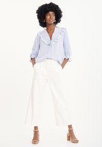 Camicia blu chiaro con colletto a volant e dettagli ricamati bianchi, abbinata a pantaloni bianchi a gamba larga e sandali con tacco quadrato color marrone chiaro.