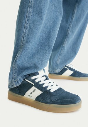 Osoba nosząca niebieskie zamszowe trampki z białymi paskami i sznurówkami, w połączeniu z luźnymi niebieskimi dżinsami na białym tle.