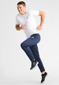 Hvid kortærmet sportstrøje med logo, parret med navy sportbukser. Modellen bærer sorte løbesko med grå detaljer.