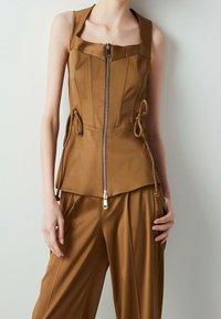 Top marrone senza maniche con chiusura frontale a zip, corpino strutturato, lacci laterali e orlo svasato, abbinato a pantaloni larghi coordinati.
